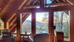 A cabin with a wooden interior, large windows, a dining table, and a view of a mountainous landscape.