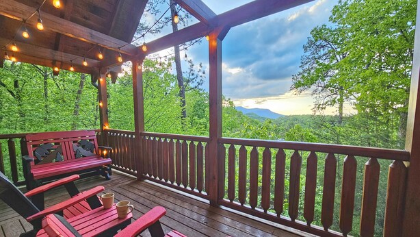 A wooden deck with red benches and string lights, overlooking a lush green forest and mountains.