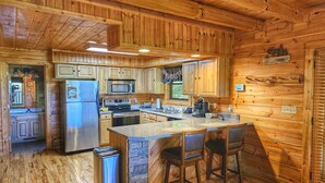 A wooden cabin interior with a kitchen featuring wooden cabinets, a refrigerator, a microwave, and a stove. There is a stone island with bar stools and a window with blinds.
