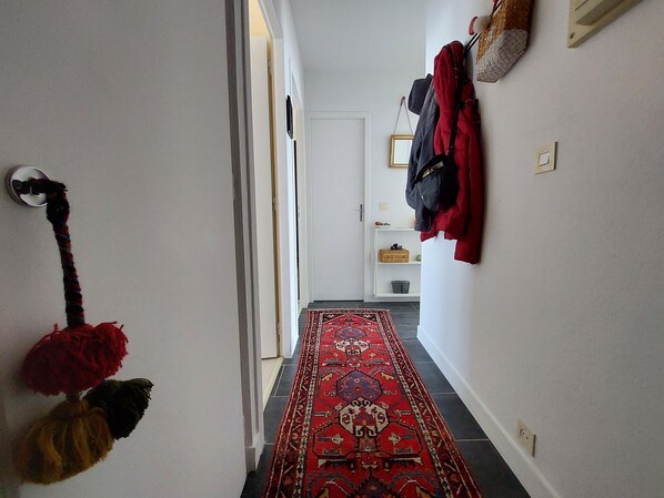 Un pasillo con un tapete rojo estampado, un perchero con abrigos y una puerta al final.