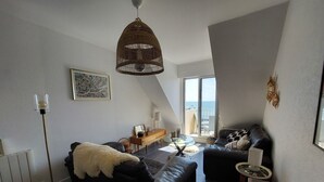 A modern living room with a black leather sofa, a white furry ottoman, a glass coffee table, and a wicker pendant light. - Wild coastline, exceptional sea view, close to shops and TGV station (Le Croisic)
