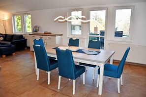 Ein moderner Essbereich mit einem weißen Tisch und blauen Stühlen, großen Fenstern mit Aussicht sowie einer Küchenzeile mit Arbeitsfläche und Geräten.