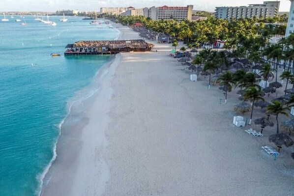 Ein Strand mit einer weiten Sandfläche, einer Pier mit Sonnenschirmen und Küstenlinie, gesäumt von Palmen.