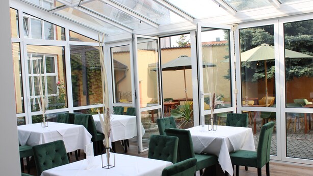 A conservatory with tables and chairs, large windows, and a view of the garden. - Spreezeit Hotel connect (Senftenberg)