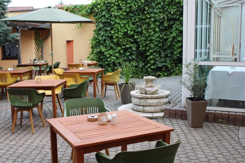 An outdoor patio area with wooden tables and chairs, a stone fountain, and a green hedge in the background.