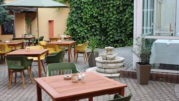 An outdoor patio area with wooden tables and chairs, a stone fountain, and a green hedge in the background.