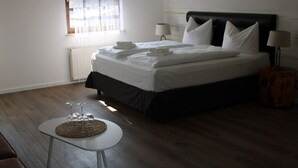 A neatly made bed with pillows and folded towels, a bedside table with a woven placemat and wine glasses, a lamp, and a window with blinds. - Spreezeit Hotel connect (Senftenberg)