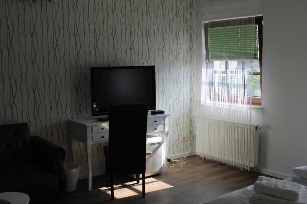 A room with a bed, a desk with a TV, a chair, a radiator, and a window with blinds.