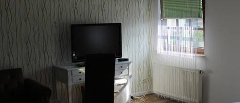 A room with a bed, a desk with a TV, a chair, a radiator, and a window with blinds.