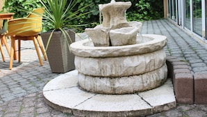 A stone fountain with a tiered design, surrounded by a paved area with a potted plant and a yellow chair. - Spreezeit Hotel connect (Senftenberg)
