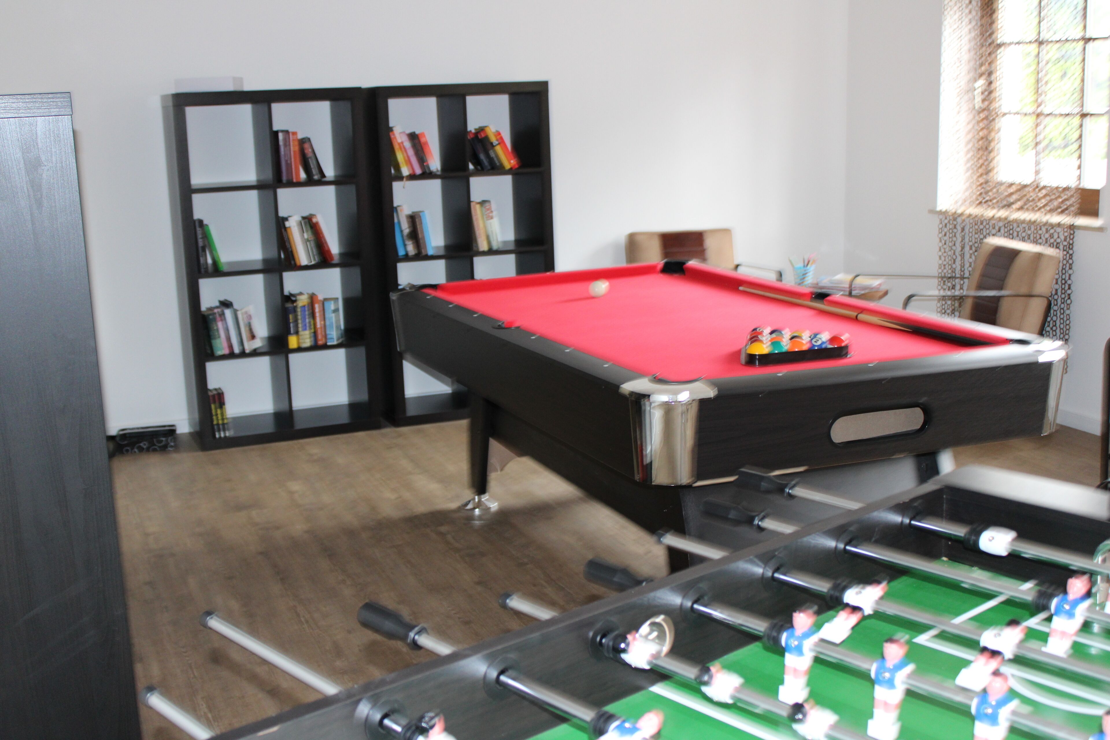 Una mesa de billar con superficie de fieltro rojo, una mesa de fútbol de mesa y una estantería llena de libros.