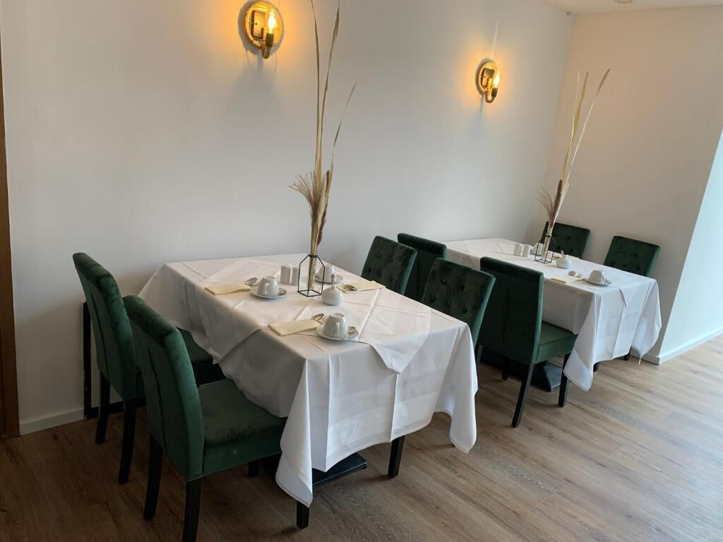 A dining area with tables set for four, each with a white tablecloth, green upholstered chairs, and decorative vases.