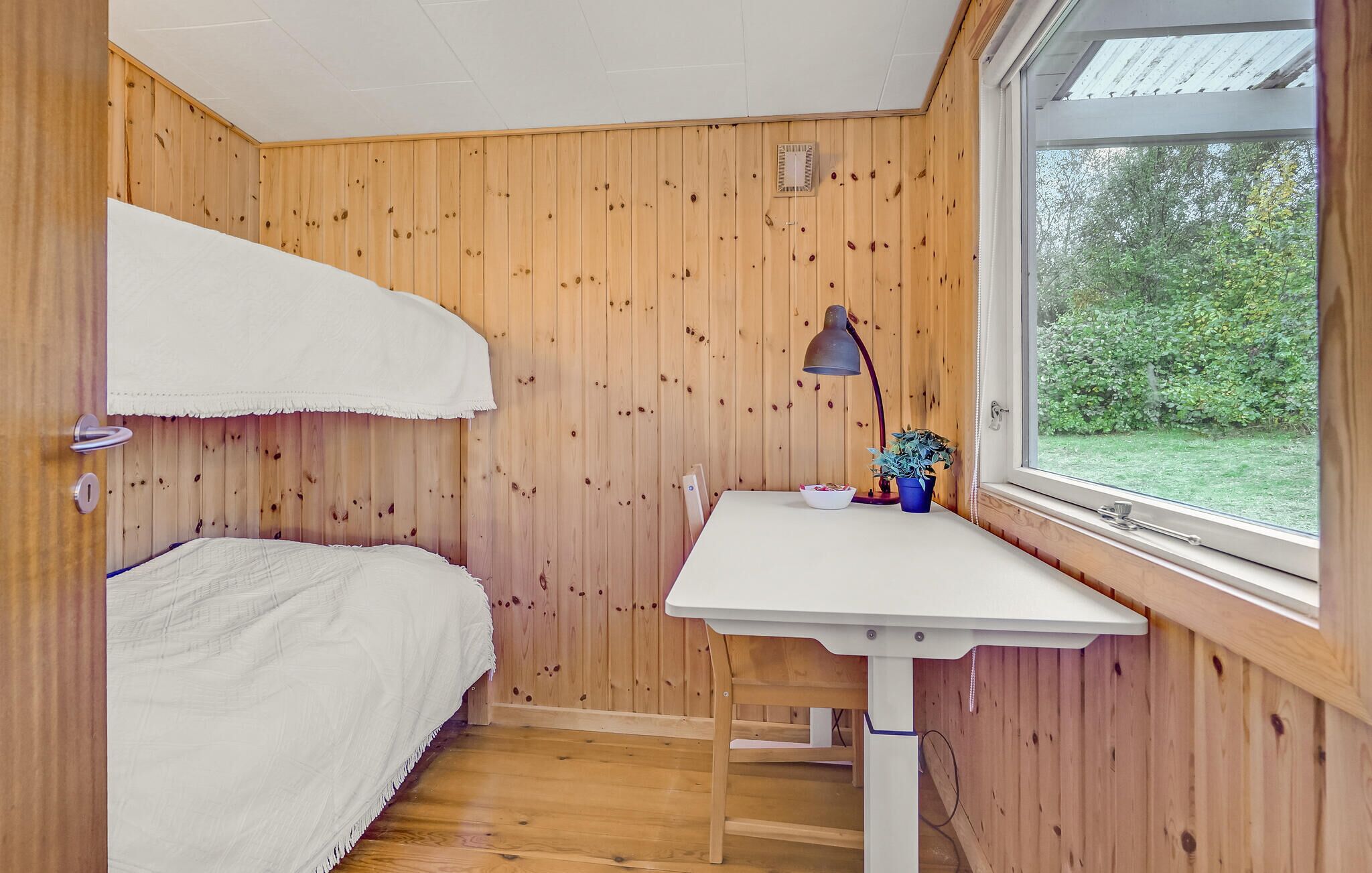 A small, cozy room with wooden walls, a bunk bed, a desk with a lamp, and a window overlooking greenery.