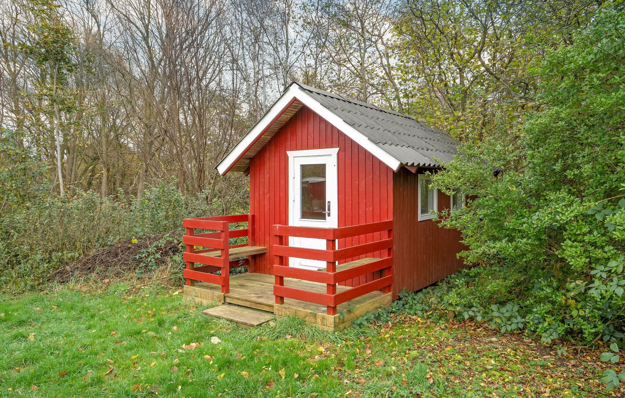 Une petite cabane en bois rouge, avec une porte et des fenêtres blanches, entourée de verdure.