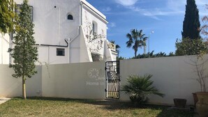 Edifici blanc amb entrada reixa, un petit jardí amb gespa i una palmera, i cel blau clar.