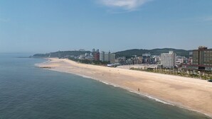 A coastal cityscape with a wide sandy beach, a clear blue sky, and a mix of modern and traditional buildings along the coastline. - Daecheon Mud Beach Hotel (Boryeong)