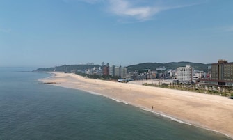 A coastal cityscape with a wide sandy beach, a clear blue sky, and a mix of modern and traditional buildings along the coastline.