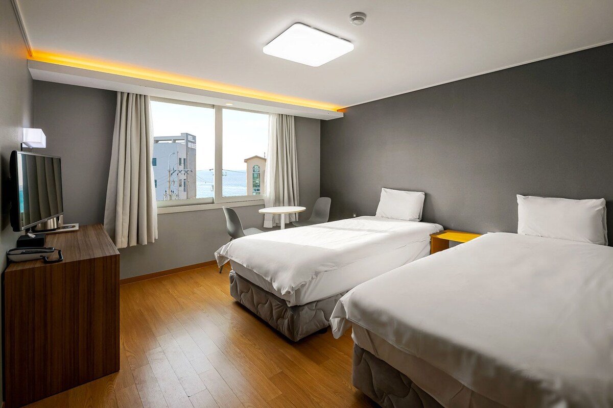 Ein Hotelzimmer mit zwei Betten, einem Fernseher, einem Schreibtisch und einem Fenster mit Blick auf Gebäude.