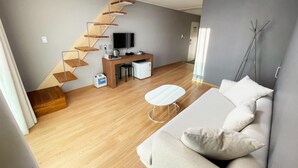 A modern living room with a wooden staircase, a white sofa, a small white table, and a TV on a wooden stand. - Pampas Hotel Jeju (Jeju City)