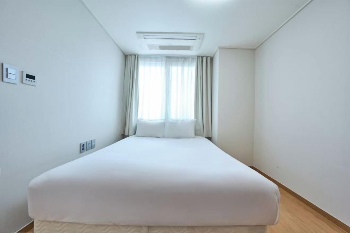Ein Hotelzimmer mit einem Einzelbett, einer Deckenklimaanlage, einem Fenster mit Vorhängen und einem an der Wand montierten Thermostat.