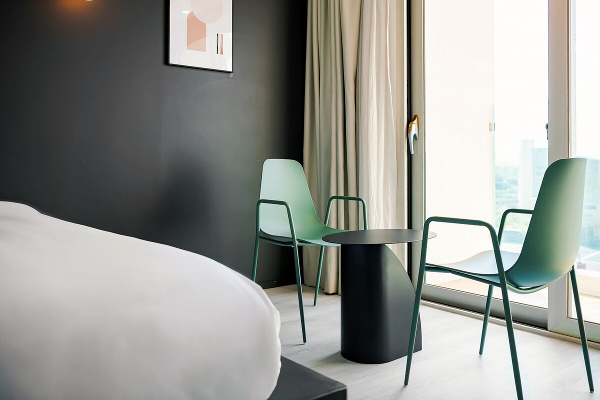 Ein modernes Hotelzimmer mit einem großen Bett, zwei Stühlen, einem kleinen Tisch und Blick auf die Stadt.