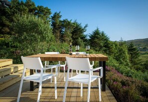 An outdoor wooden deck with a table and chairs, surrounded by greenery and hills.