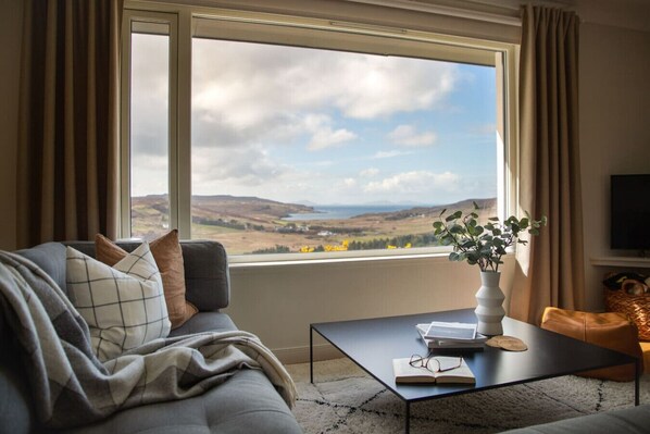 Ein Wohnzimmer mit einer Couch, einem Couchtisch, einer Pflanze und Blick auf die Landschaft.