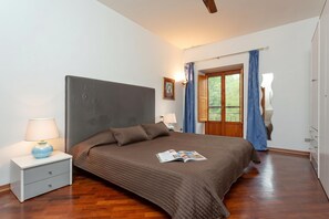 Een slaapkamer met een groot bed, een nachtkastje met een lamp, een raam met gordijnen en een houten vloer.
