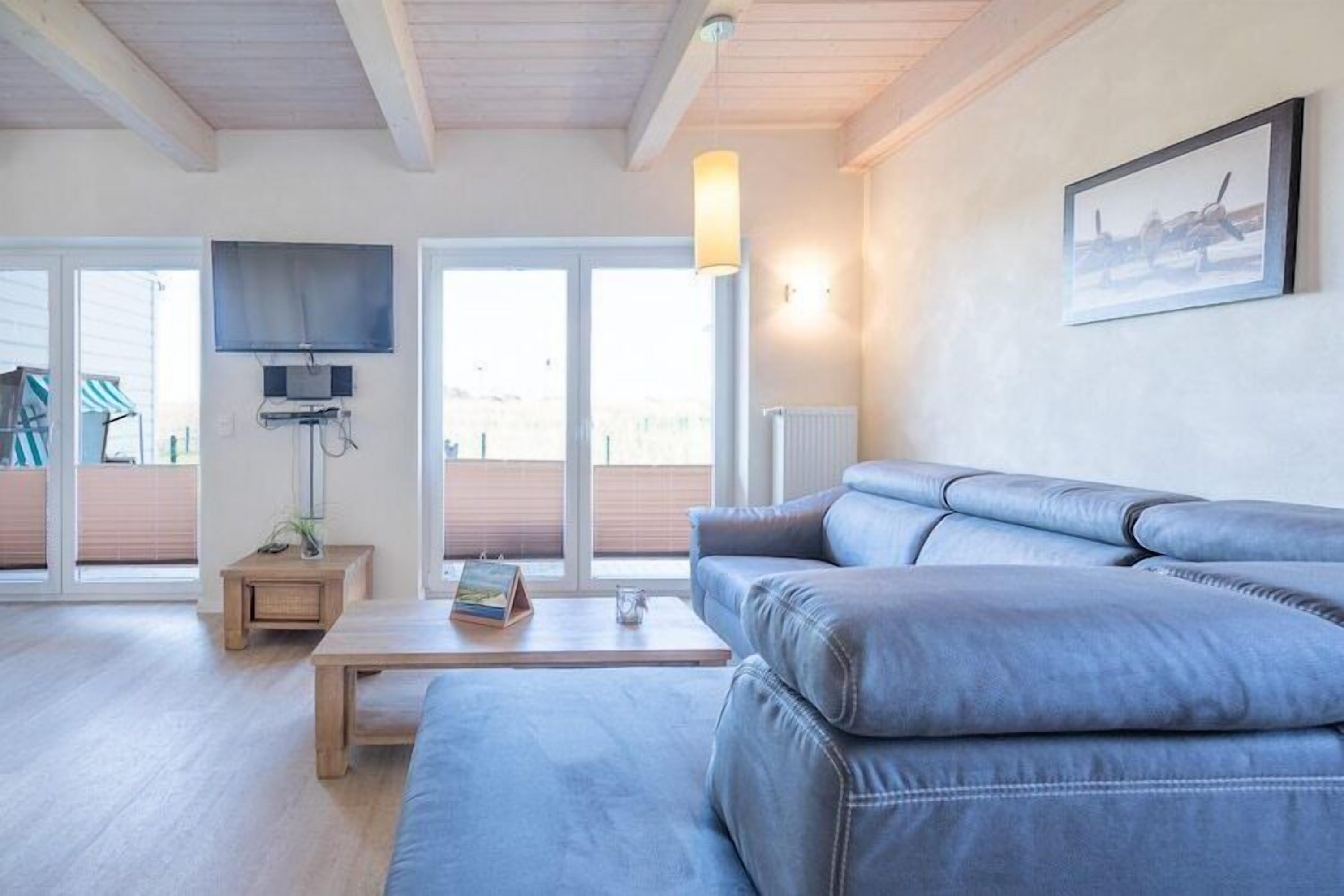 Ein modernes Wohnzimmer mit einer großen grauen Couch, einem Holztisch, einem flachbildfernseher an der Wand und einem großen Fenster mit Blick auf eine Holzterrasse.