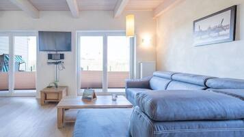 Ein modernes Wohnzimmer mit einer großen grauen Couch, einem Holztisch, einem flachbildfernseher an der Wand und einem großen Fenster mit Blick auf eine Holzterrasse.