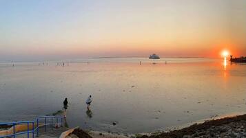 Ein Strand bei Sonnenuntergang mit einem Schiff in der Ferne, Menschen, die entlanggehen, und einer Treppe, die zum Wasser führt.