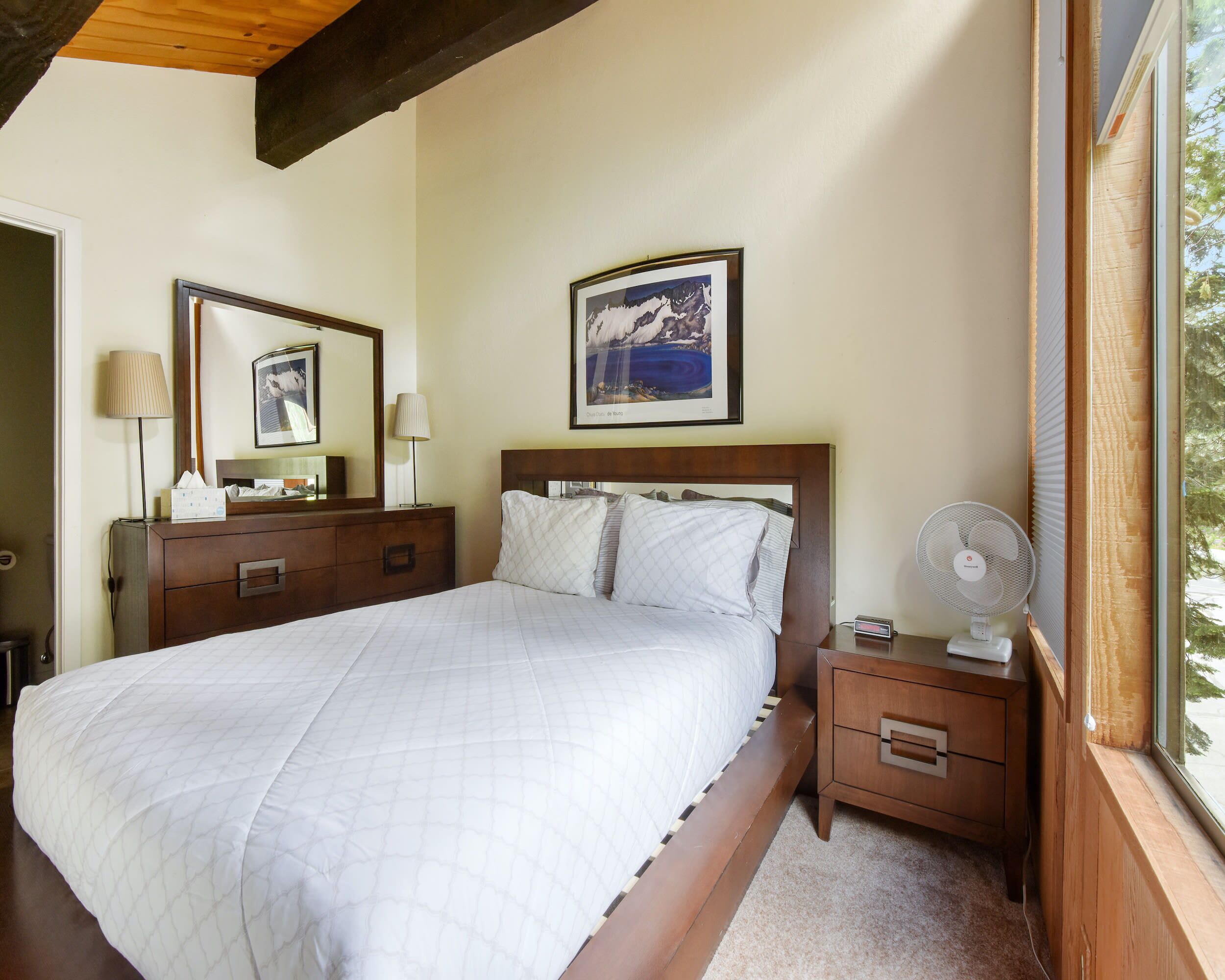 Cama bem arrumada com colcha branca acolchoada, cabeceira de madeira e mesa de cabeceira com abajur e ventilador. Uma moldura com quadro está pendurada acima da cama, e uma janela com persianas é visível à direita.