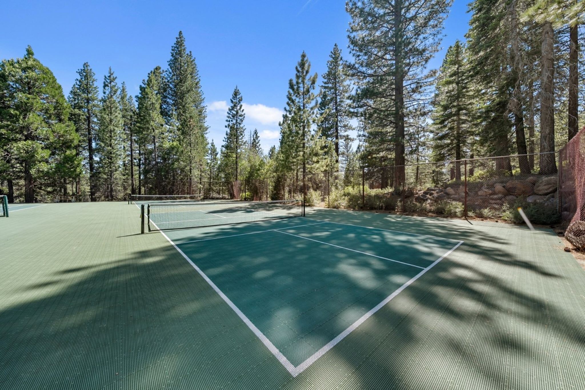 An outdoor tennis court surrounded by tall trees and a rocky outcrop.