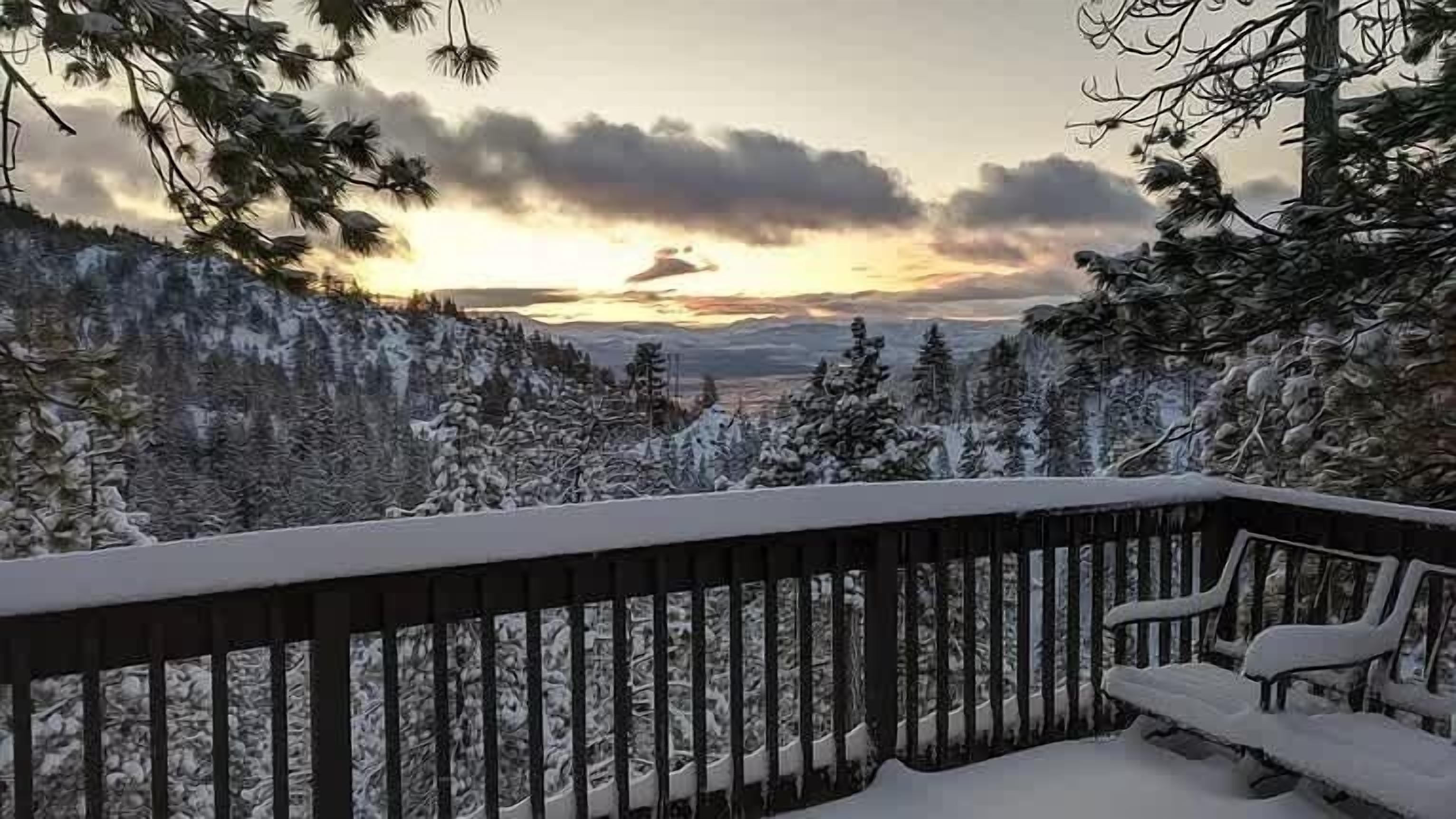 A snow-covered deck with a view of a forest and mountains during sunset.