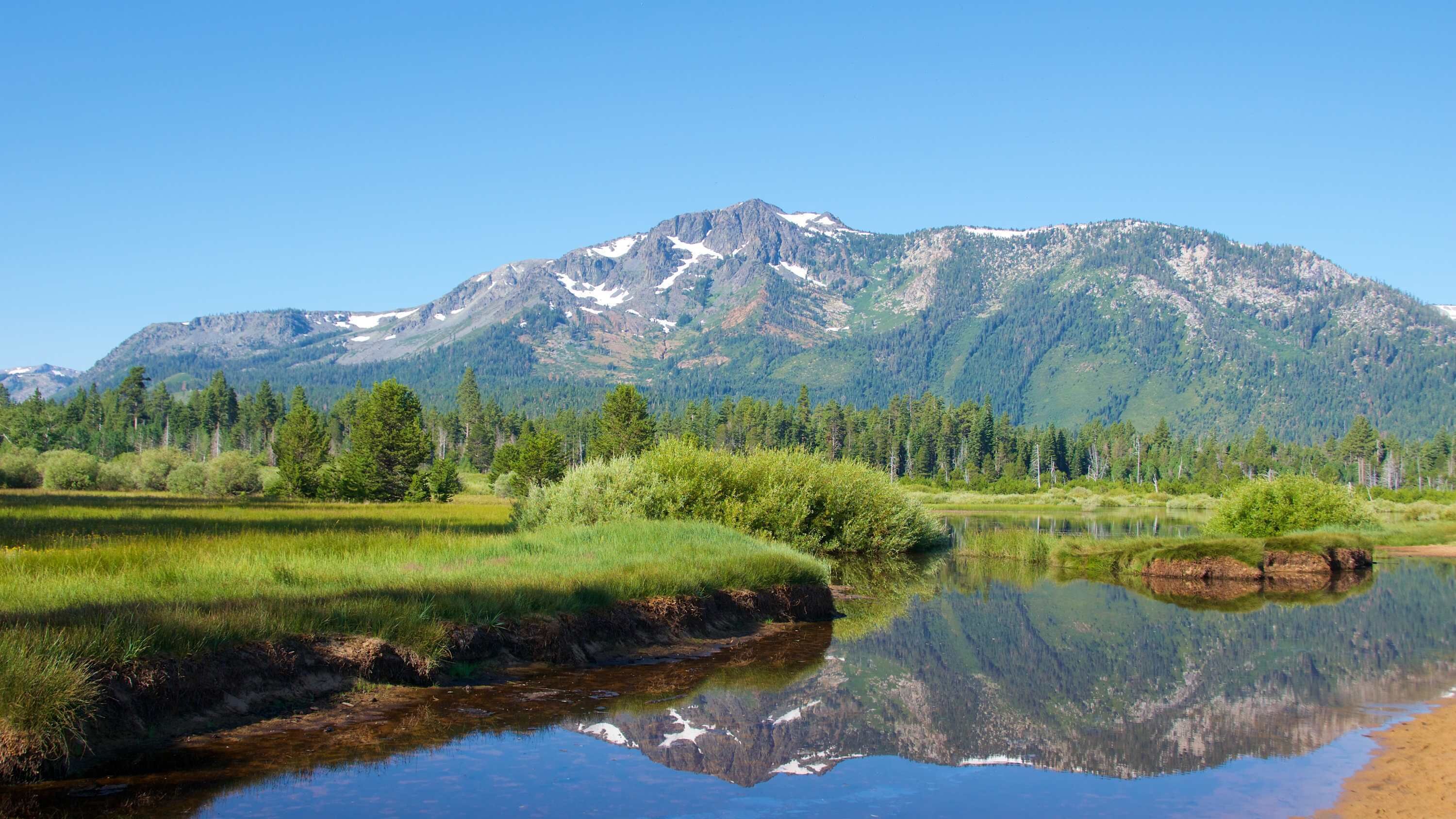 A mountain landscape with a clear reflection in a calm body of water, surrounded by dense forest and grassy areas.