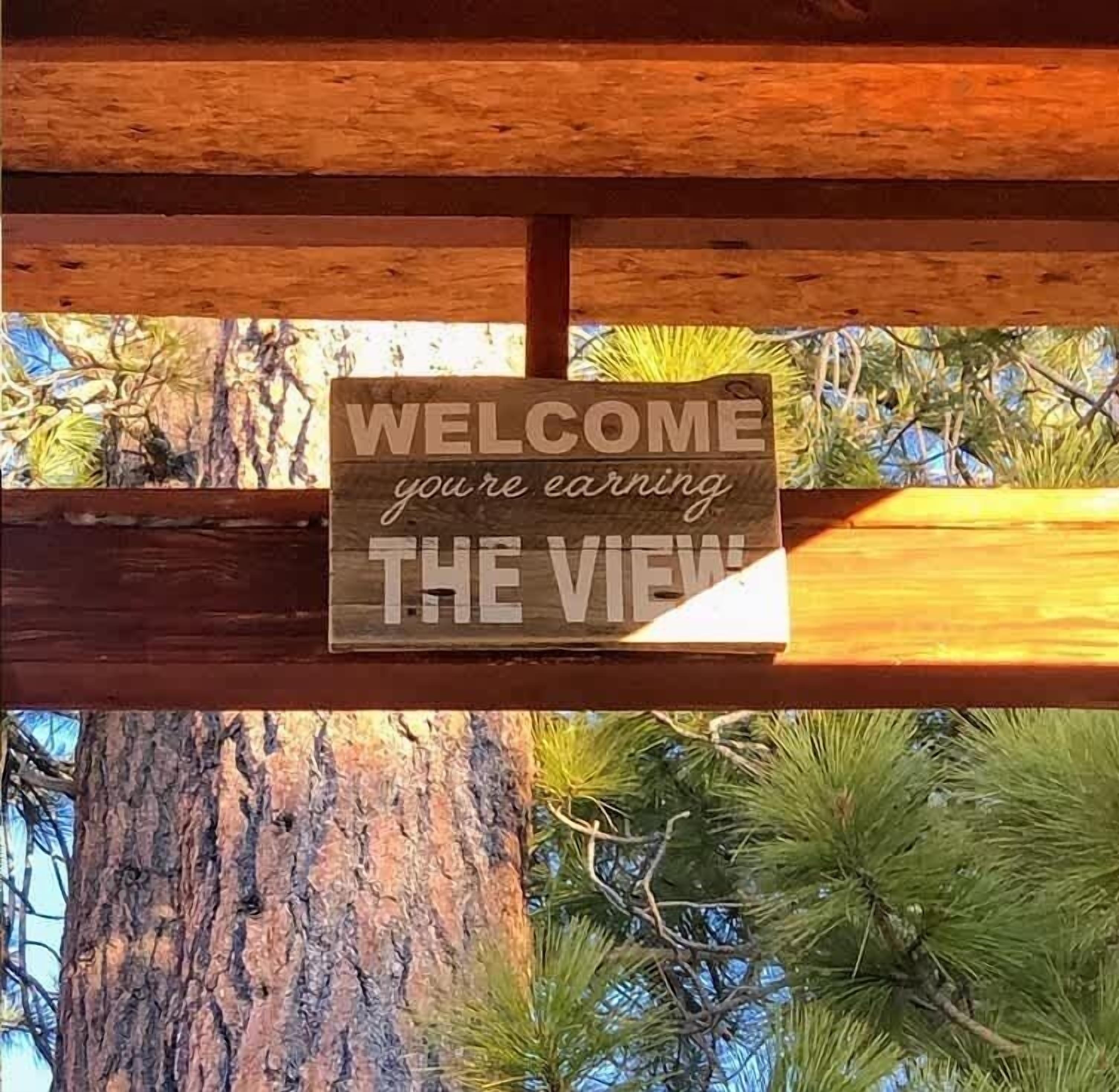 Ein hölzernes Schild mit der Aufschrift „WELCOME you're earning THE VIEW“ vor einem Baum und blauem Himmel.