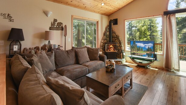 A cozy living room with a stone fireplace, a large window with a scenic view, and a wooden ceiling. - Lux Leisure Game Room & More! (Truckee)