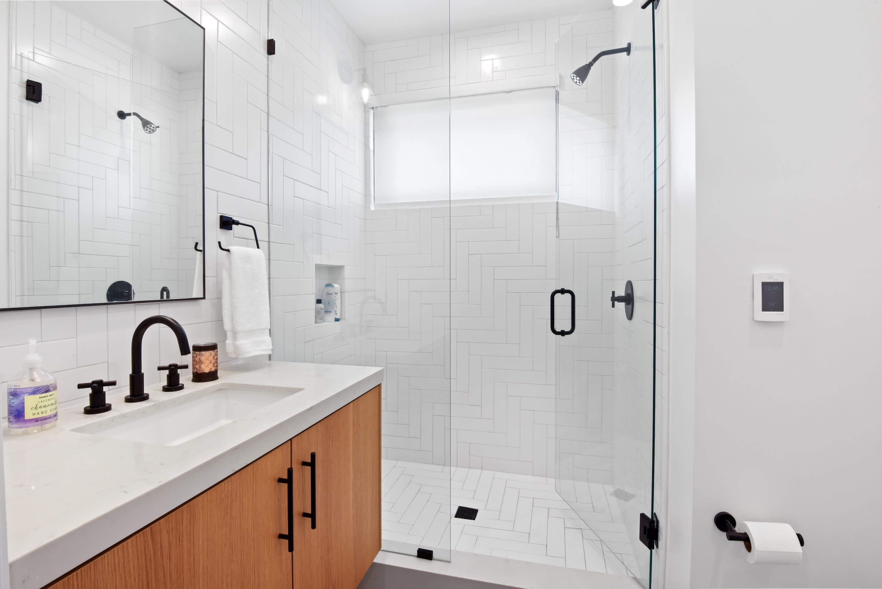 A modern bathroom with a glass-enclosed shower, a white tiled wall, a wooden vanity, and a large mirror.
