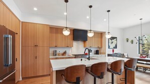 A modern kitchen with wooden cabinets, a central island with bar stools, stainless steel appliances, pendant lighting, and a view of the outdoors. - Enjoy Paradise Hot Tub, Bike & Beach Equipment! (Olympic Valley)