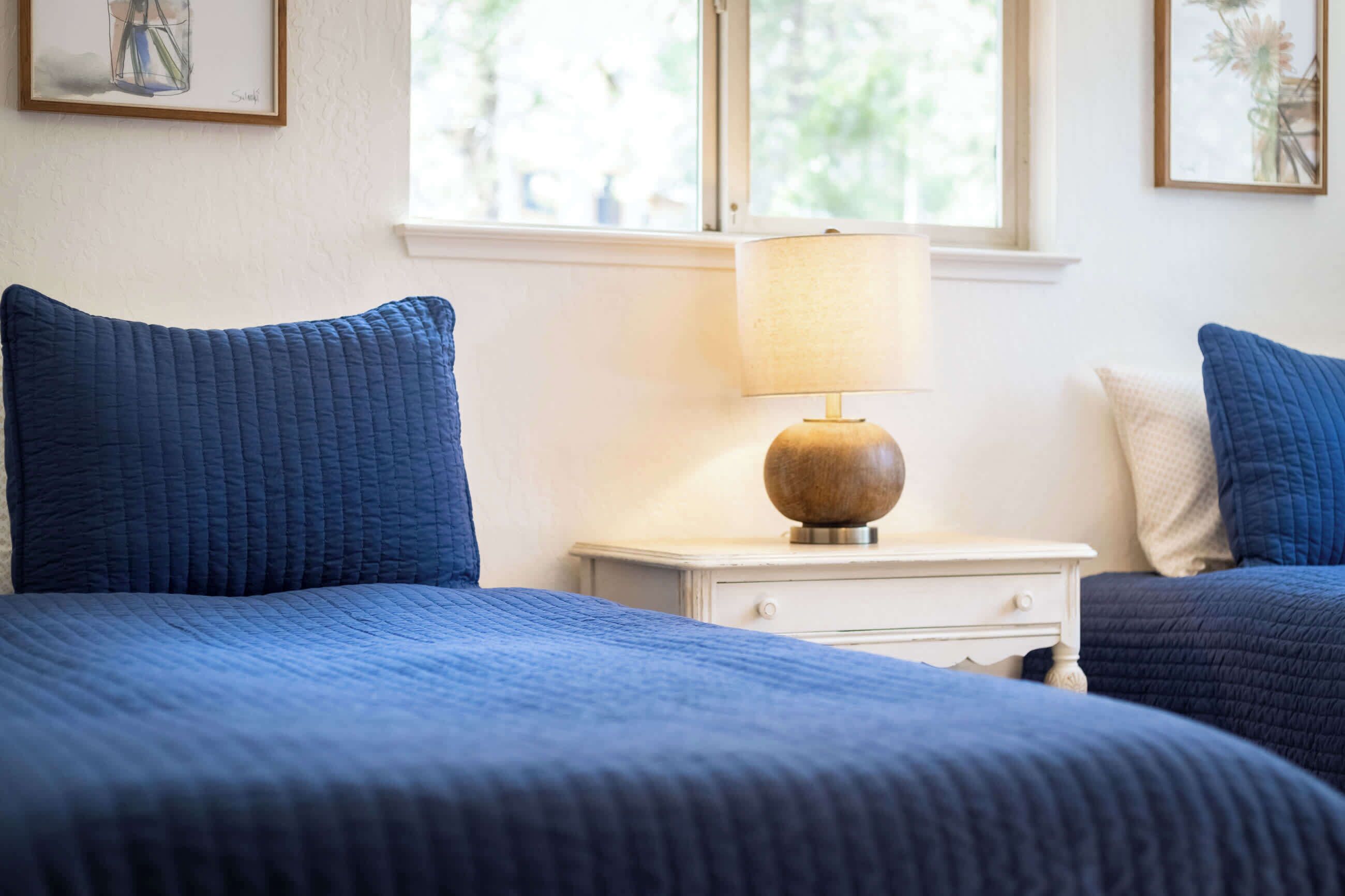 A bedroom with two beds, a bedside table with a lamp, and a window with a view of greenery.