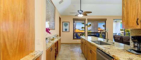A kitchen with wooden cabinets, a granite countertop, a ceiling fan, and large windows overlooking a pool area.