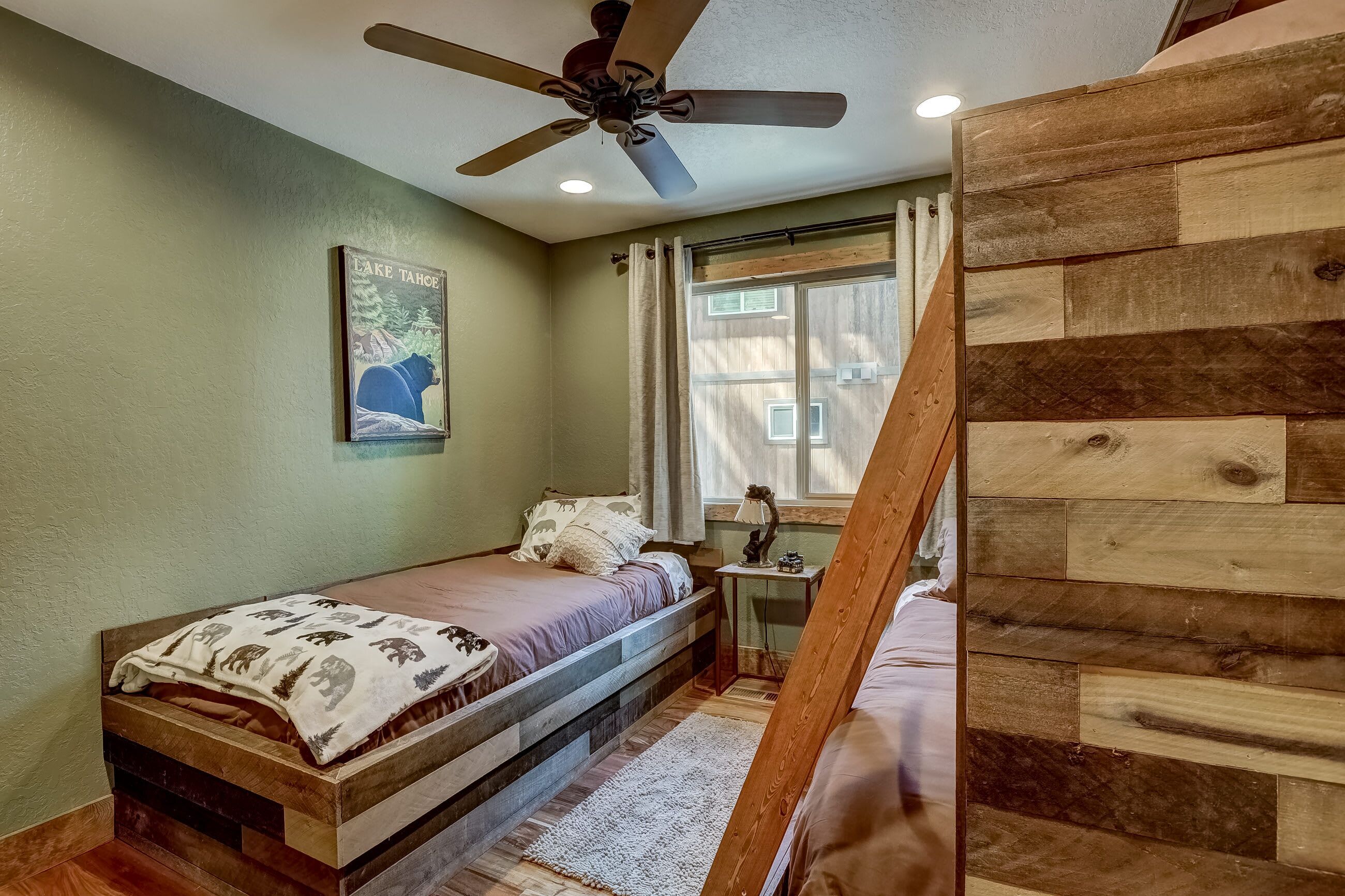 Une chambre avec un lit en bois, un ventilateur de plafond, une table de chevet avec une lampe, une fenêtre avec des rideaux et une affiche accrochée au mur.