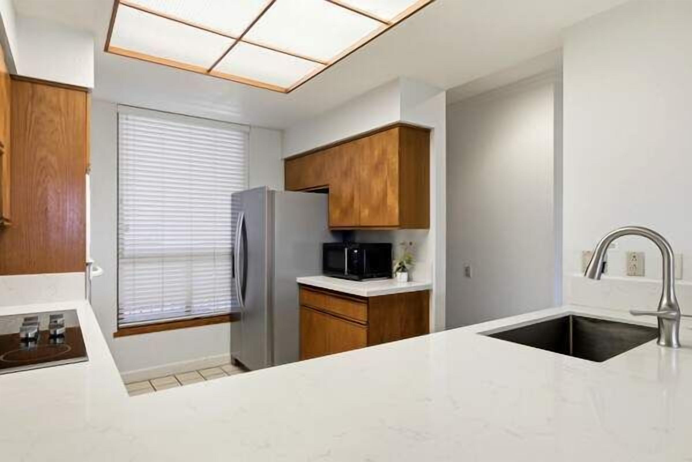 A modern kitchen with white countertops, wooden cabinets, a stainless steel refrigerator, a microwave, and a sink with a faucet.