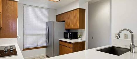 A modern kitchen with white countertops, wooden cabinets, a stainless steel refrigerator, a microwave, and a sink with a faucet.