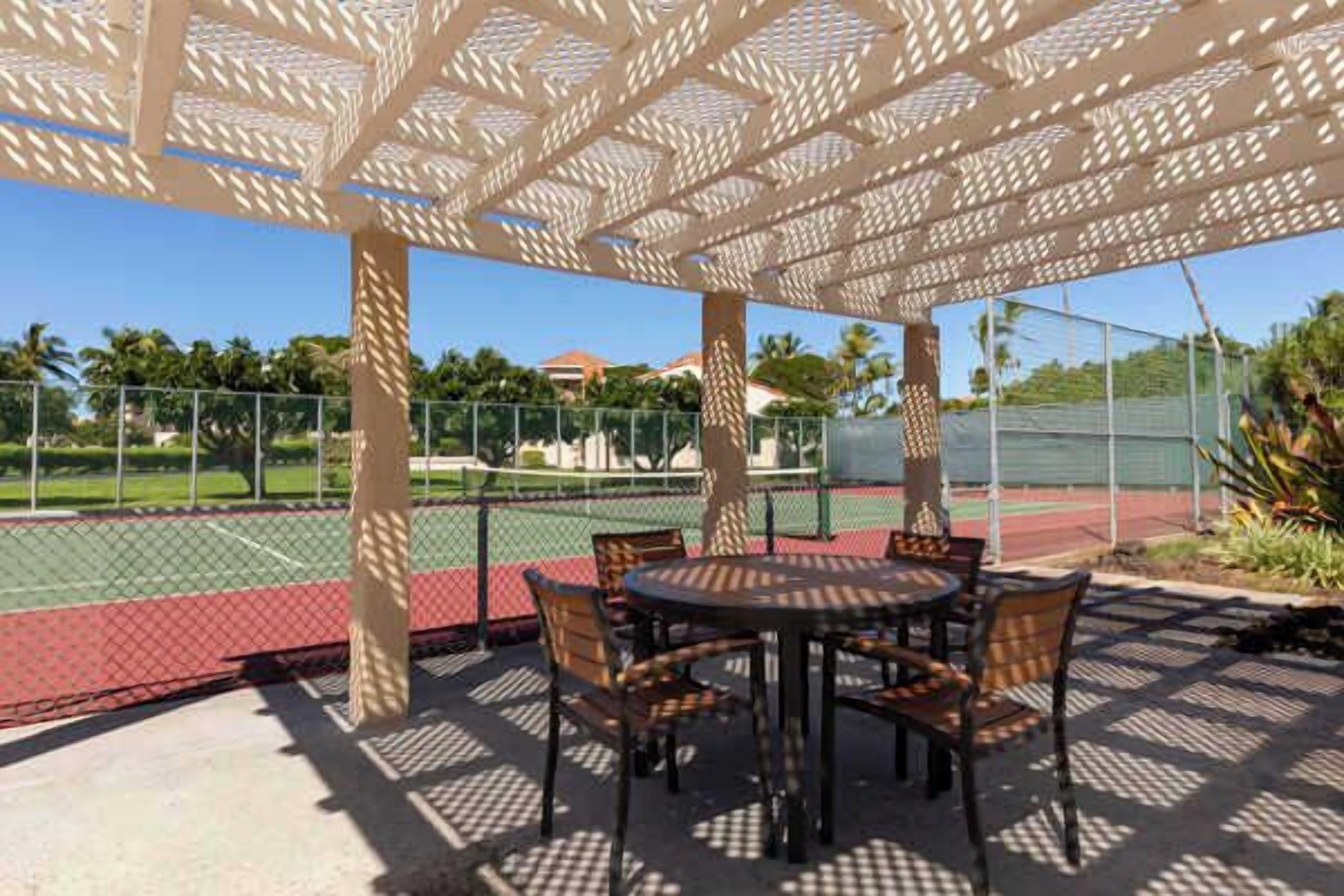 An outdoor patio with a table and chairs, a tennis court in the background, and a patterned canopy overhead.