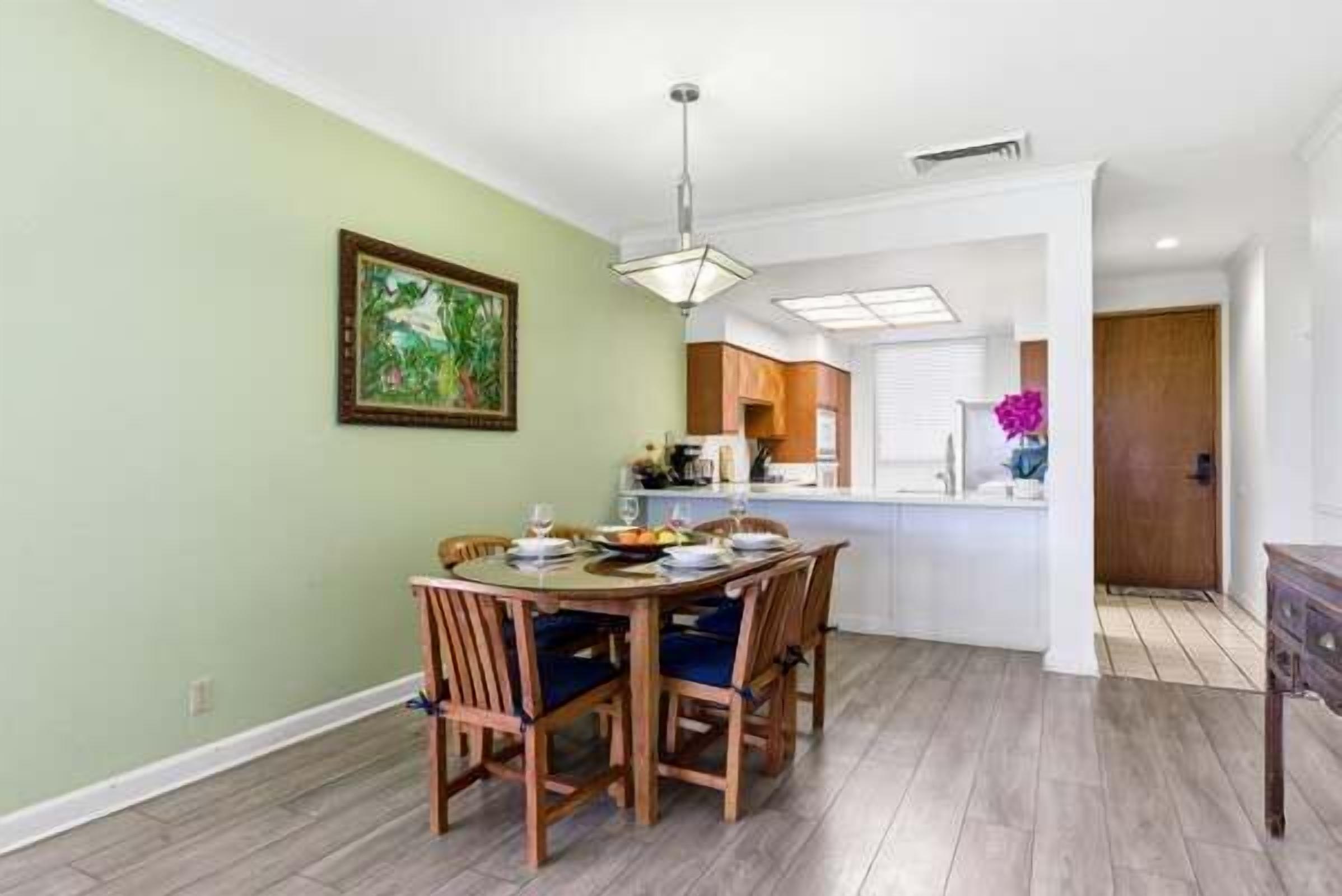 A dining area with a wooden table and chairs, a painting on the wall, and a kitchen in the background.