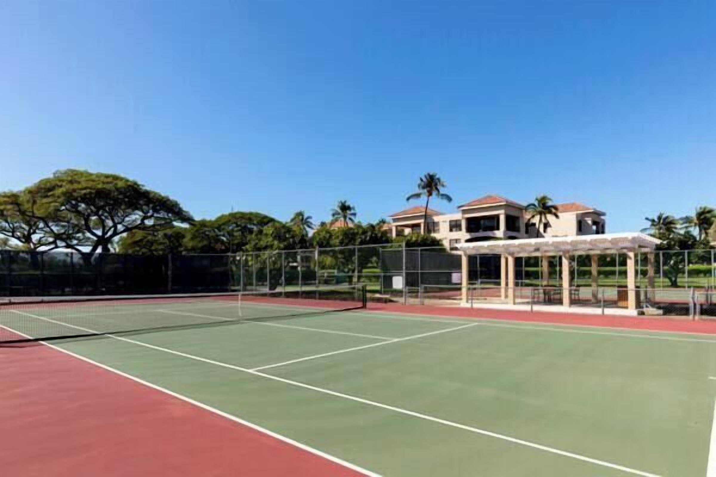 An outdoor tennis court with a net, surrounded by a fence, and buildings in the background.