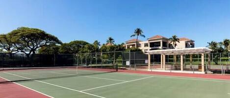 An outdoor tennis court with a net, surrounded by a fence, and buildings in the background.