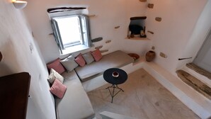 A high-ceilinged room with a stone wall, a window, a sofa with cushions, a small round table, and a wooden bench. - Kastro Cozy Guesthouse (Paros)