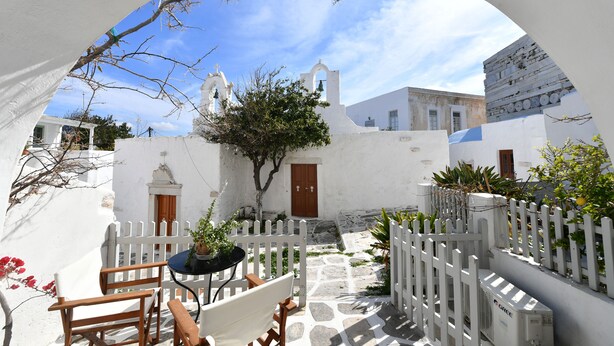 A courtyard with a white fence, a table with chairs, and a building with a bell tower. - Kastro Cozy Guesthouse (Paros)